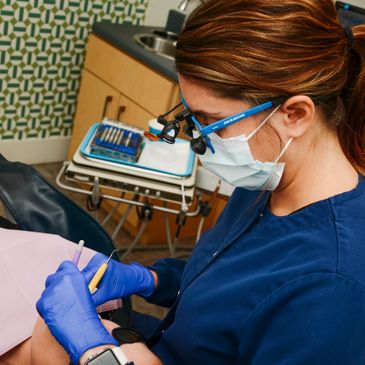 Dentist wearing blue gloves and magnifying glasses performing dental work.