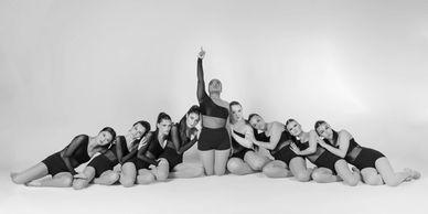 A group of dancers in black outfits posing on the floor with one standing and pointing upward.