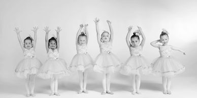 Six young ballerinas in peach tutus posing with raised arms against a white background.