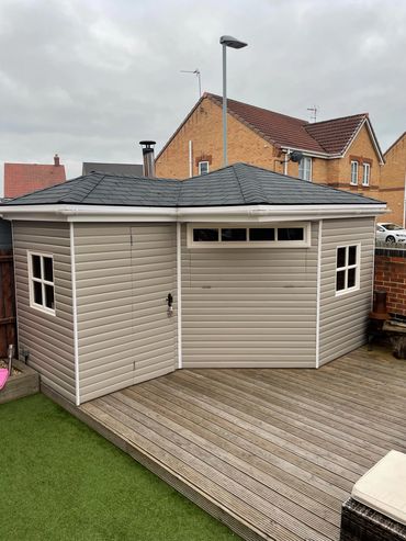 A modern beige garden shed with windows on a wooden deck beside artificial grass.