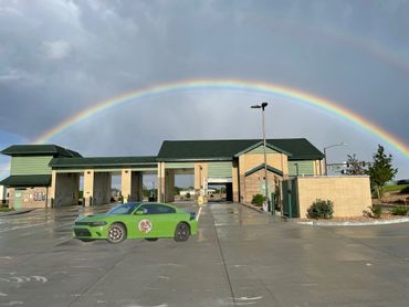 A green car with a dog logo parked near a building under a vibrant rainbow.