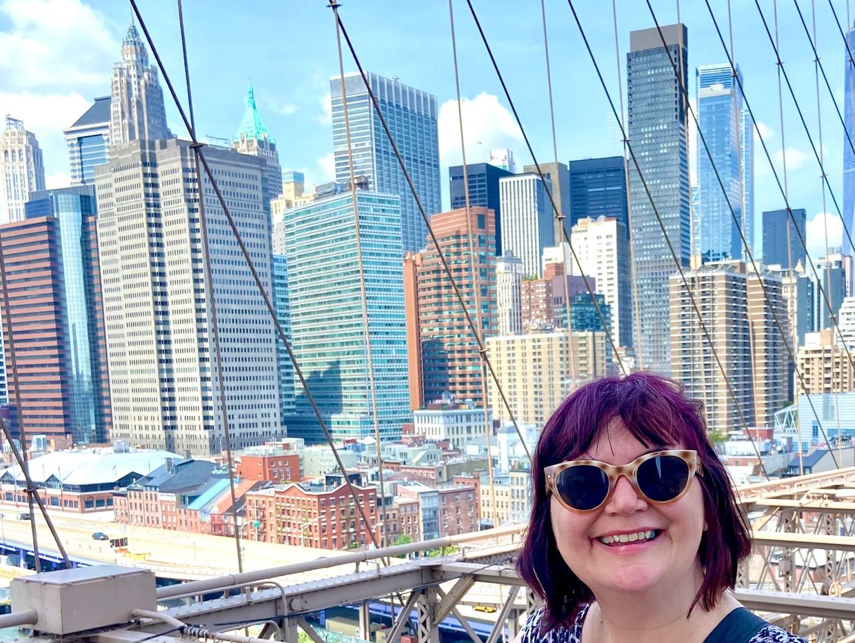 Smiling woman with sunglasses on a bridge, city skyline in the background.