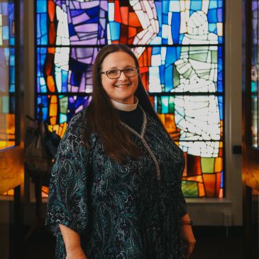 Woman wearing clerical collar standing in front of a stained glass window.