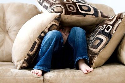 Child in denim jeans sitting on couch hiding under pillows