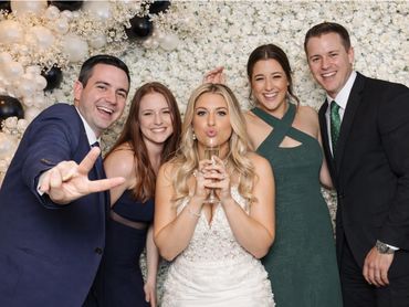 Five friends joyfully pose at a celebration with a floral and balloon backdrop.
