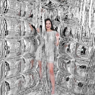 A woman joyfully posing inside a shiny silver balloon room with metallic streamers.