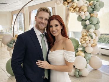 A happy couple dressed elegantly poses in front of a decorative balloon arch indoors.