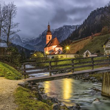 Lit church by a stream with mountains in the background at dusk.