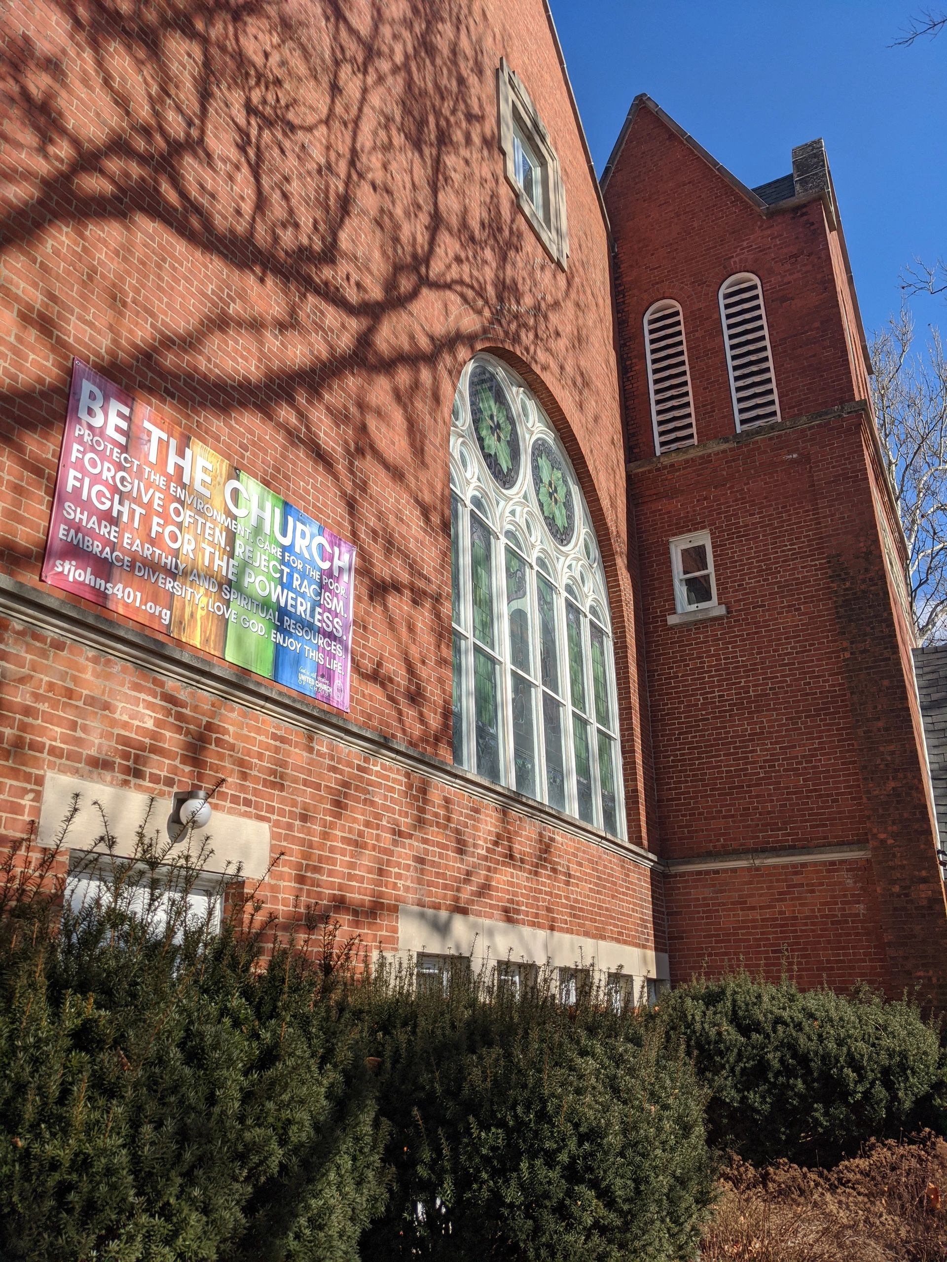Front of church building with rainbow banner reading "Be The Church" and stained glass window.