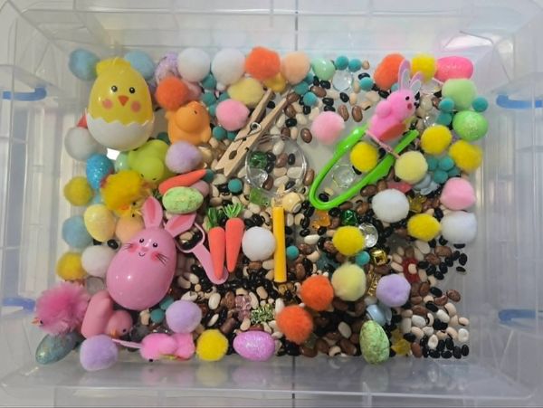 A sensory bin filled with colorful pom-poms, plastic eggs, small toys, and beads for tactile play.