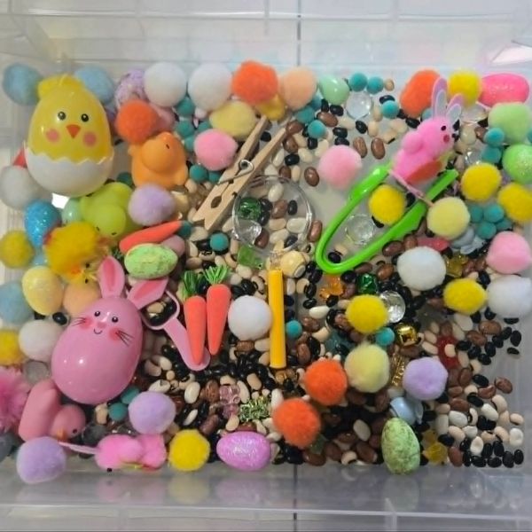 A sensory bin filled with colorful pom-poms, plastic eggs, small toys, and beads for tactile play.