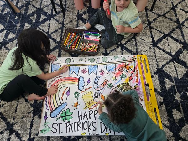 Three children coloring a St. Patrick's Day poster with markers and crayons.