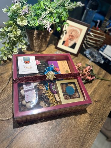 Diwali gift box with sweets and potpourri on a wooden table beside flowers and a framed photo.