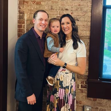 A smiling family of three posing indoors against a brick wall.