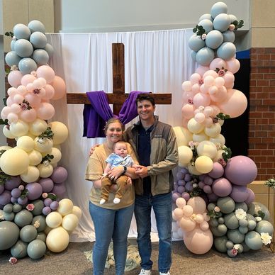 A smiling family of three posing in front of a wooden cross and pastel balloon decorations.