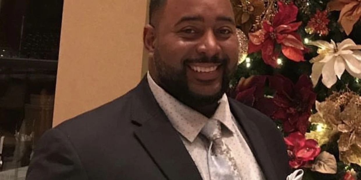 Man in a suit smiling beside a decorated Christmas tree.