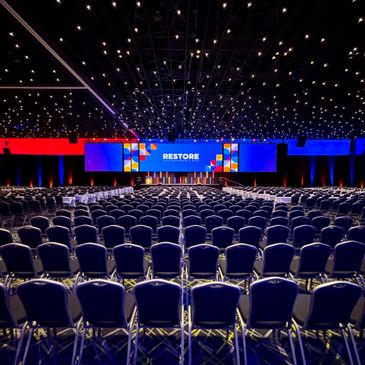 Empty conference hall with blue chairs facing a stage labeled RESTORE.