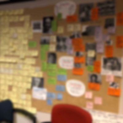 Blurred image of a wall covered with sticky notes and photos, with chairs in front.