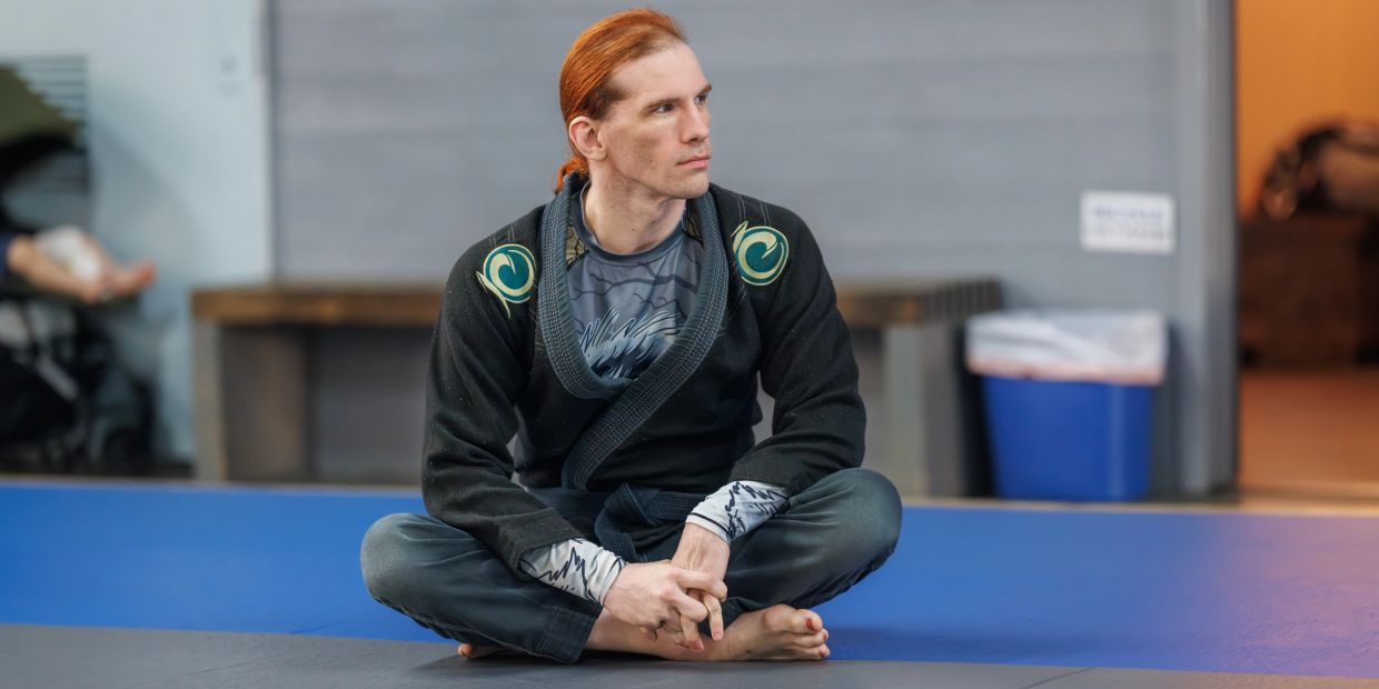 Man in martial arts gi sitting cross-legged on mat, looking contemplative.