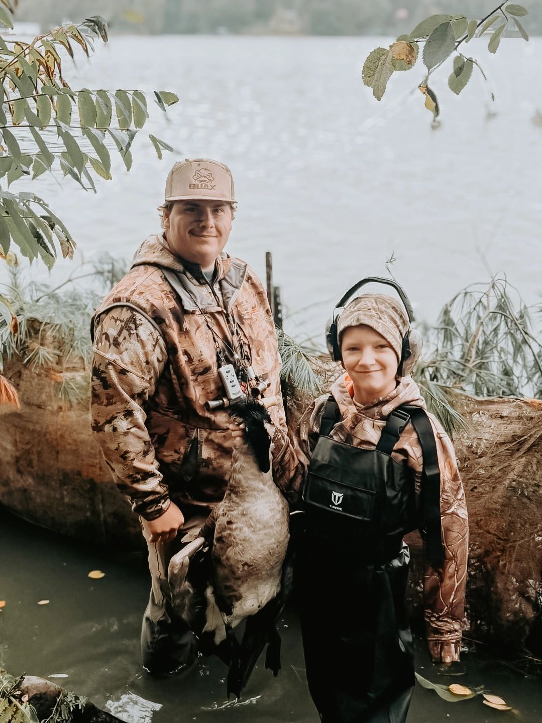 Two hunters posing with a harvested goose near water.