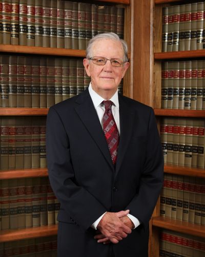 Elderly man in suit standing in front of a bookshelf filled with legal books.