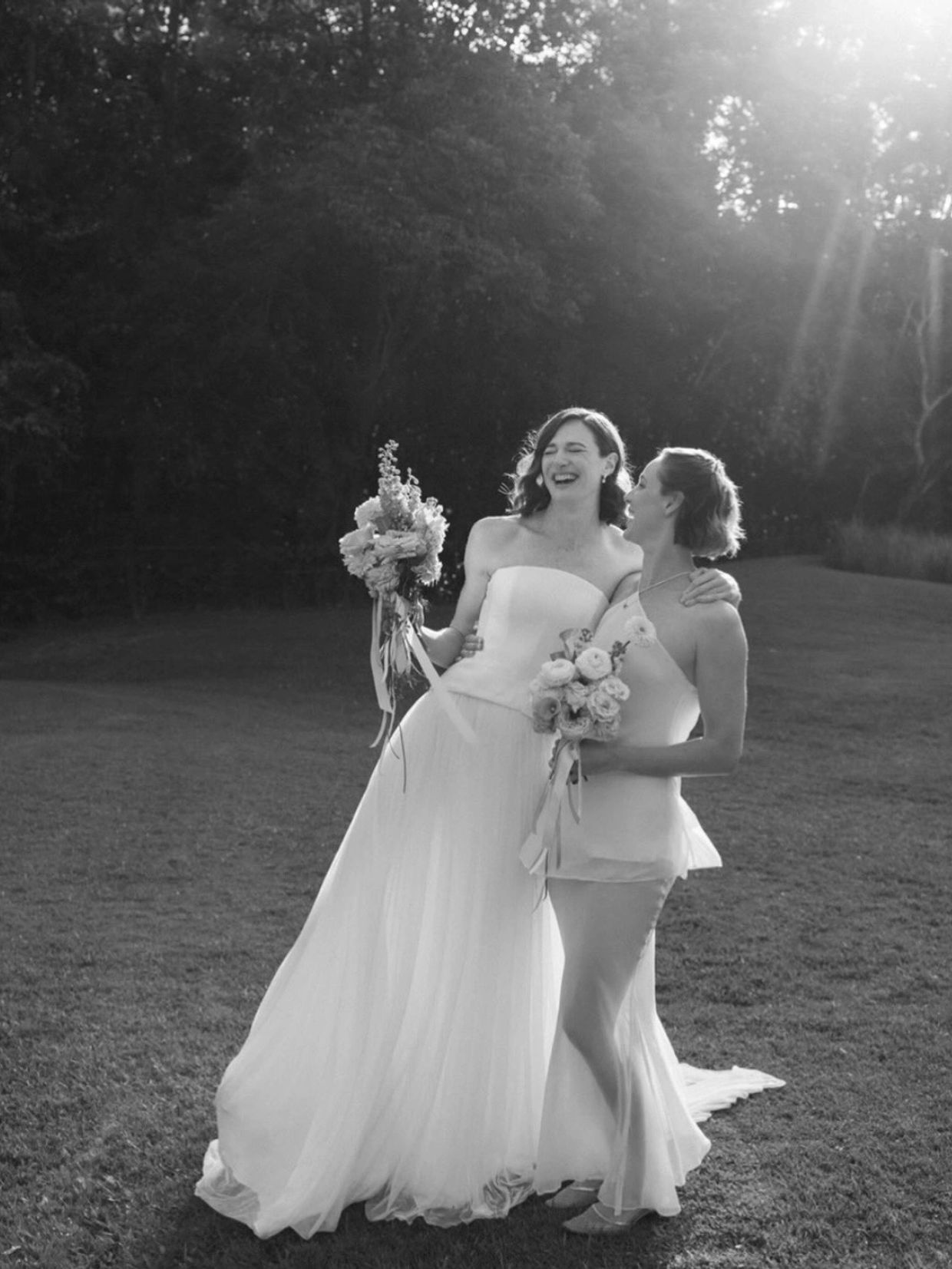 Two brides smiling and holding bouquets outdoors.