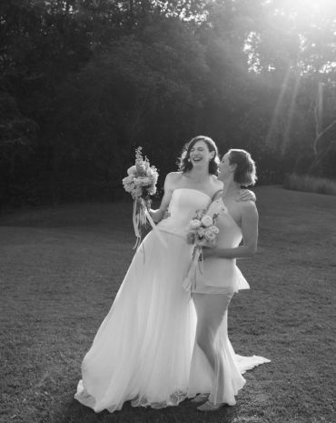 Two brides smiling and holding bouquets outdoors.