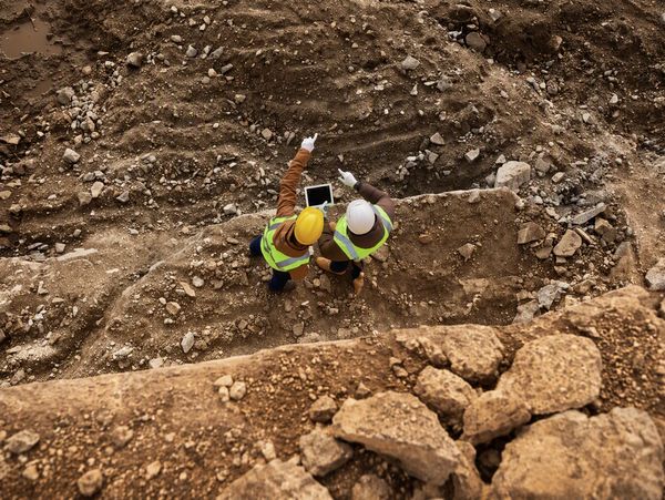 Two construction workers in safety gear inspecting a dirt site with a tablet.