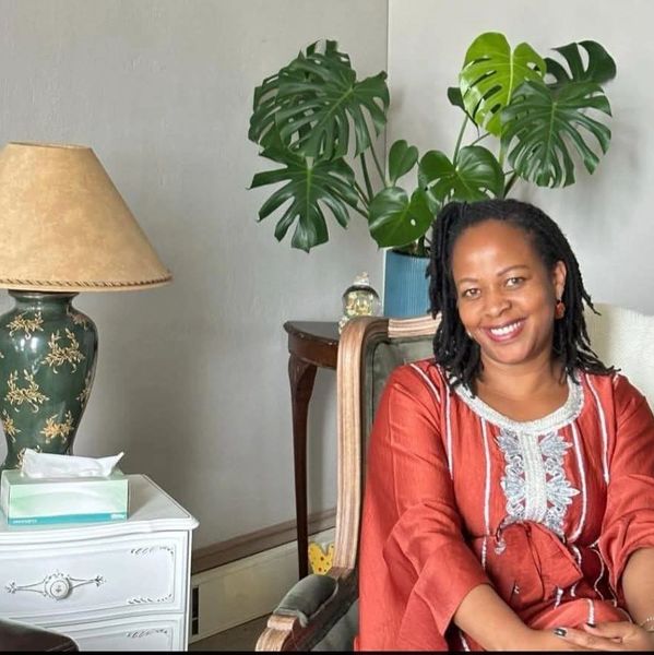 Smiling woman seated indoors with a plant and lamp in the background.