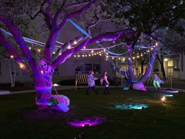 Two actors stand in between the trees and under fairy lights and streamers. It is night time.