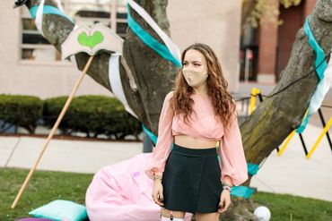 An actor stands in front of a tree, bean bag, and pillows. A totem of hands rests in the background