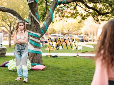 An actor stands in front of a tree full of streamers with their arms crossed. They're talking.