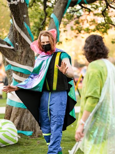 An actor stands in front of a tree full of streamers and addresses another actor.