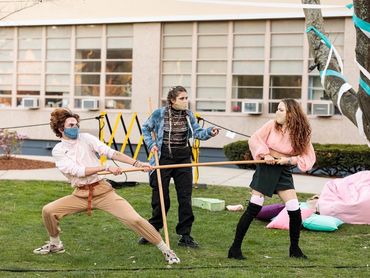 Two actors tug on the ends of a large stick, fighting with each other. A third stands in between.