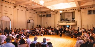 A ballroom full of people sitting in a circle. House lights are on.