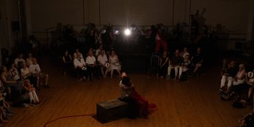 A single bulb shines above an actor holding another actor, sitting on a crate center stage.