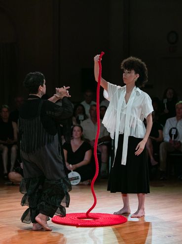 An actor picks up the end of a red rope coil. She faces another actor.