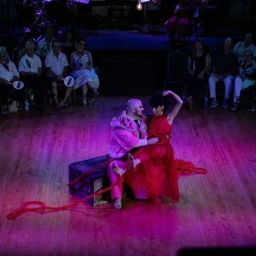 Two actors sit on a single crate in the middle of the circular space. They are playful.