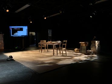 The stage is empty of actors. On it are a table, three chairs, and a variety of boxes upstage left.