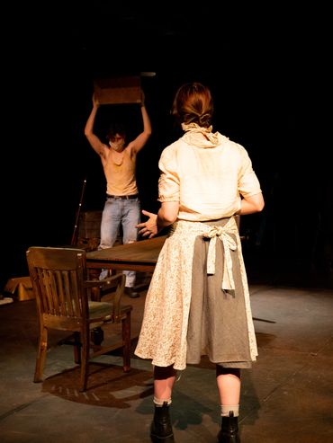 In the foreground, an actor is turned away towards another actor holding a box above his head.