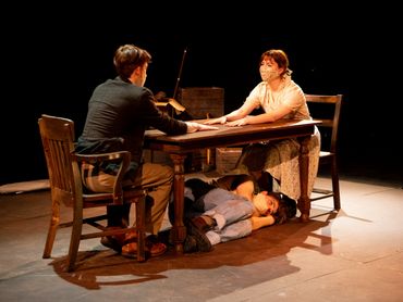 Two actors sit across from each other at a table. A third actor lays on the floor under the table.