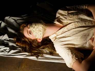 An actor lays on the floor on top of a sheet. She looks towards a bright light.