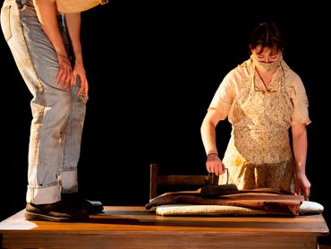 An actor stands on a table, while another actor irons fabric.