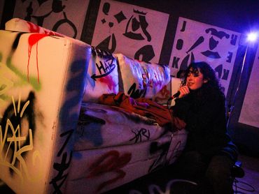 An actor kneels and leans onto a spray painted couch. A wall is covered in large playing cards.
