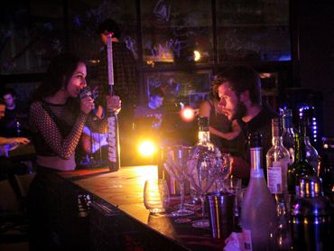 An actor sings into a mic holding a baseball bat. Another actor sits at the bar drinking.