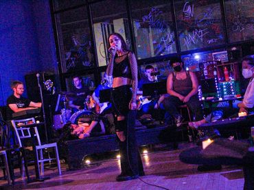 An actor sings into a wired hand held microphone. Behind them, the band sits on a raised platform.