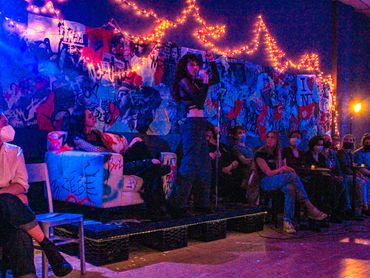 Two actors are on a platform in front of the mural. One sits and watches, another sings into mic.
