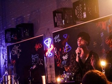 An actor sings with mic. Behind are mirrors with graffiti and floating milk crate shelves.