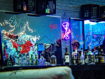 A view of a bar with bottles scattered. Behind, a mirror is covered in graffiti inspired script.