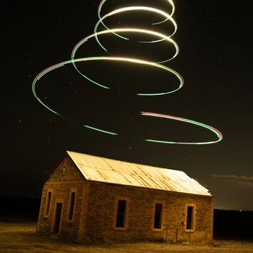 Light trails spiral above a rustic stone building at night under a starry sky.